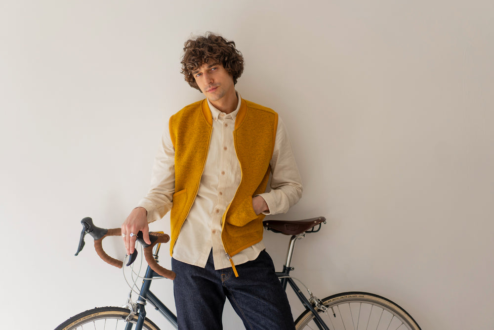 Man wearing a mustard yellow vest standing next to a bicycle against a plain background